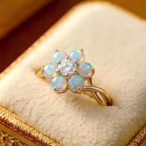 May include: A gold ring featuring a flower-shaped cluster of six round cabochon opals surrounding a single central round brilliant-cut diamond. The band has a split, twisted design that connects to the base of the floral setting.