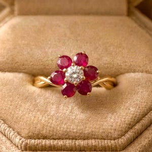 Könnte beinhalten: Ein Goldring mit einem zentralen runden weißen Diamanten, der von sechs tiefroten Rubinen in Blütenform umgeben ist. Die Ringschiene ist gedreht gestaltet und die Fassung besteht aus poliertem Gelbgold, was ein Blumenmotiv ergibt.