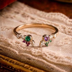 Puede incluir: Un anillo abierto de oro con dos puntos focales. Un lado muestra una piedra preciosa redonda verde acentuada por pequeñas piedras rojas y claras. El otro lado presenta una piedra preciosa redonda morada con pequeñas piedras verdes y claras.