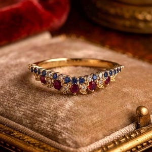 May include: A gold curved ring featuring a row of small round blue sapphires and red rubies alternating with tiny clear diamonds. The band has a delicate metal leaf-like texture surrounding each gemstone, creating a continuous arc design.