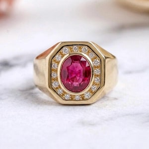 Può includere: Un anello in oro con una pietra preziosa centrale dal taglio ovale di un vibrante colore rosso rosato, circondata da un alone di piccoli diamanti bianchi rotondi. La montatura è un anello in oro lucido con una cornice ottagonale centrale.