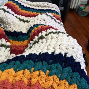 May include: A chunky knit blanket in a rainbow of colors, including cream, red, orange, yellow, teal, and navy blue. The blanket has a textured, plush appearance, perfect for adding warmth and style to any space.