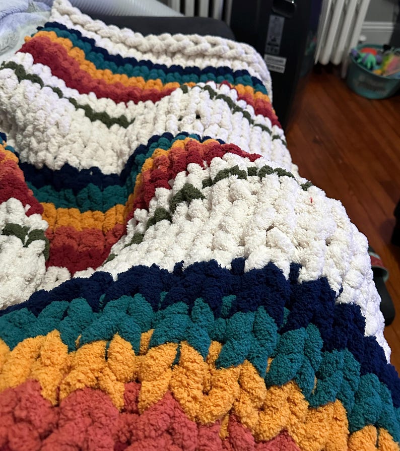 May include: A chunky knit blanket in a rainbow of colors, including cream, red, orange, yellow, teal, and navy blue. The blanket has a textured, plush appearance, perfect for adding warmth and style to any space.