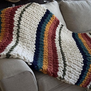 May include: A chunky knit blanket in cream, red, orange, teal, and navy blue stripes is draped over a gray sofa. The blanket has a thick, textured appearance, with a soft, inviting look. The color palette is warm and inviting.