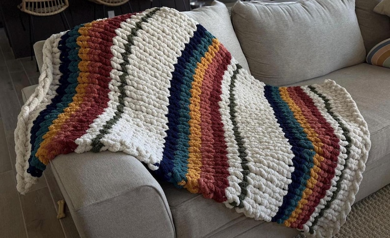 May include: A chunky knit blanket in cream, red, orange, teal, and navy blue stripes is draped over a gray sofa. The blanket has a thick, textured appearance, with a soft, inviting look. The color palette is warm and inviting.