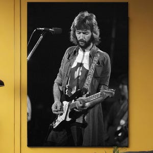 Puede incluir: Una impresión enmarcada en blanco y negro de un músico tocando la guitarra en el escenario. La imagen se muestra sobre un sofá amarillo en un salón. La habitación también cuenta con un sillón gris, una mesa de centro negra y una ventana grande.