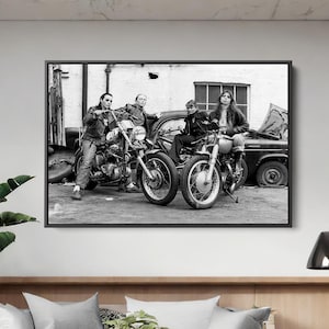 May include: Black and white framed photograph featuring four people posing with two motorcycles and a vintage car. The image is a medium shot, capturing the subjects in a garage setting. The motorcycles are parked in front of the car.