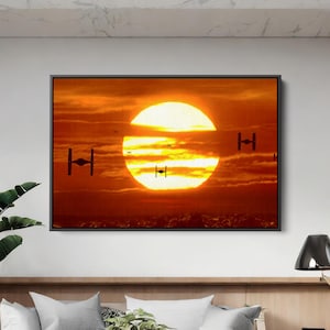 May include: Framed artwork depicting a sunset with a large, bright sun in the center. Silhouetted TIE fighters and other spacecraft are visible against the orange and yellow sky. The artwork is displayed above a white sofa.