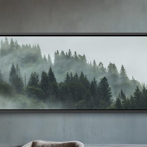 Puede incluir: Una escena de salón con una gran obra de arte paisajística enmarcada que representa un bosque brumoso. Dos sillones grises se enfrentan, con una pequeña mesa de madera entre ellos. Una lámpara colgante dorada cuelga encima y una gran planta en maceta está a la izquierda.