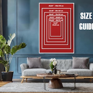 Puede incluir: Un cuadro rojo y blanco enmarcado con una gu&iacute;a de tallas, con medidas en cent&iacute;metros y pulgadas. El cuadro se muestra en una pared azul sobre un sof&aacute; gris. El texto "SIZE GUIDE" es blanco.