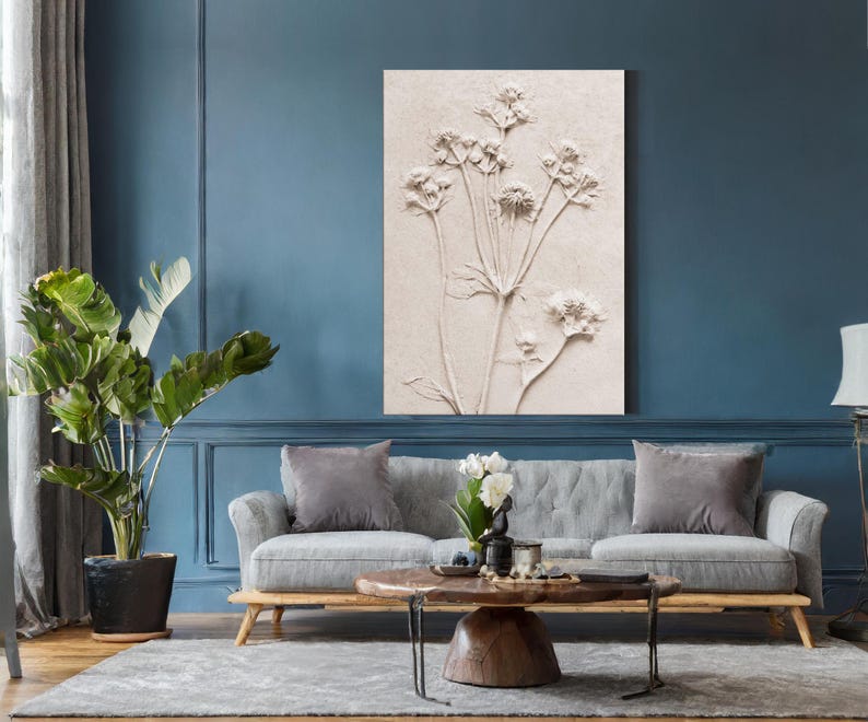 Puede incluir: Una obra de arte en relieve floral beige cuelga sobre un sof&aacute; gris. La obra de arte presenta una representaci&oacute;n detallada de varias flores con largos tallos. La habitaci&oacute;n tiene paredes azules, una alfombra gris y una gran planta en maceta.