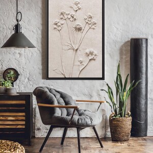 Puede incluir: Obra de arte en relieve bot&aacute;nico enmarcada en un marco negro. Presenta una representaci&oacute;n beige de una planta con flores. La habitaci&oacute;n incluye una silla gris con mechones, una cesta tejida y una planta alta en maceta.