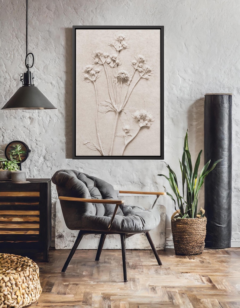 Puede incluir: Obra de arte en relieve bot&aacute;nico enmarcada en un marco negro. Presenta una representaci&oacute;n beige de una planta con flores. La habitaci&oacute;n incluye una silla gris con mechones, una cesta tejida y una planta alta en maceta.