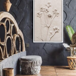 Puede incluir: Una obra de arte en relieve enmarcada, de color beige, con un dise&ntilde;o bot&aacute;nico de delicadas flores y tallos. La obra de arte est&aacute; colocada contra una pared oscura y texturizada. La habitaci&oacute;n tambi&eacute;n incluye un otom&aacute;n gris con flecos y muebles de madera.