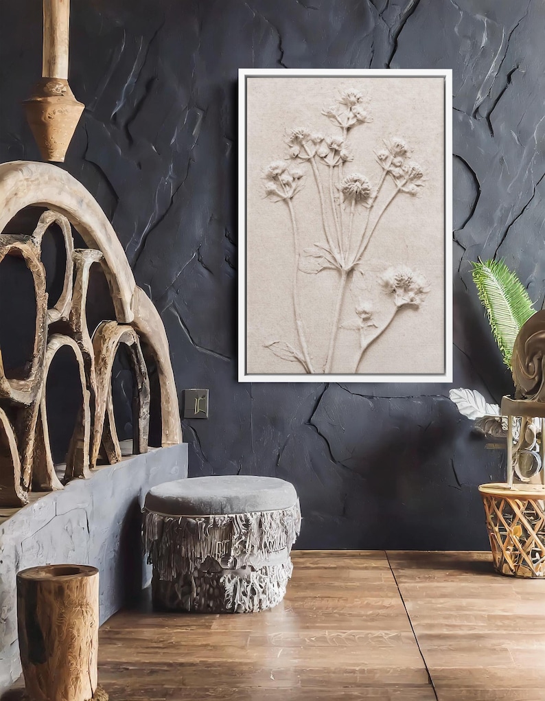 Puede incluir: Una obra de arte en relieve enmarcada, de color beige, con un dise&ntilde;o bot&aacute;nico de delicadas flores y tallos. La obra de arte est&aacute; colocada contra una pared oscura y texturizada. La habitaci&oacute;n tambi&eacute;n incluye un otom&aacute;n gris con flecos y muebles de madera.