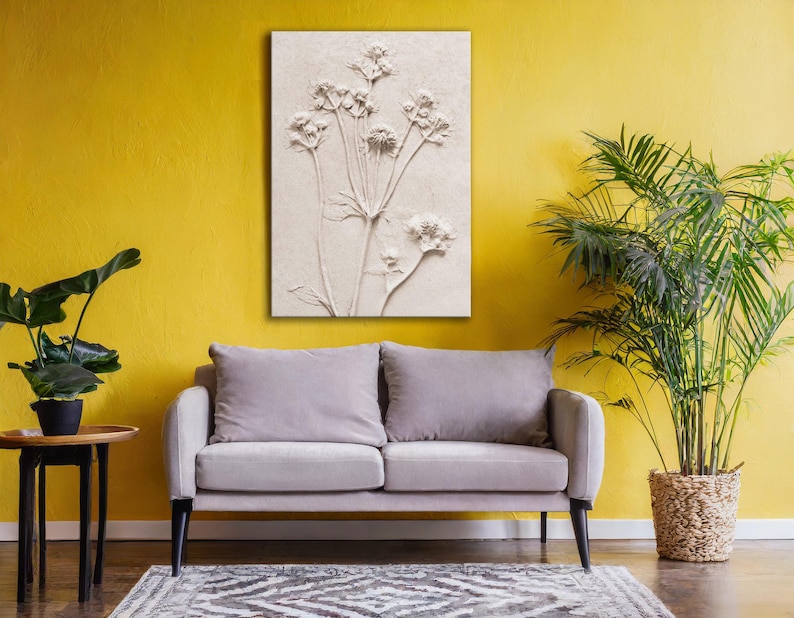Puede incluir: Una obra de arte floral en relieve de tonos neutros cuelga sobre un sof&aacute; gris. La obra de arte presenta una representaci&oacute;n detallada de flores con tallos y hojas. La habitaci&oacute;n tiene una pared amarilla, una alfombra estampada y una planta en maceta.