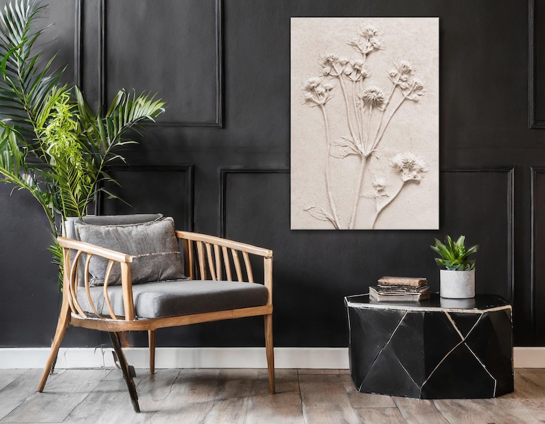 Puede incluir: Una obra de arte floral en relieve de tonos neutros cuelga de una pared negra. Una silla de madera con cojines grises y una peque&ntilde;a mesa auxiliar negra con una planta y libros completan la decoraci&oacute;n de la habitaci&oacute;n.