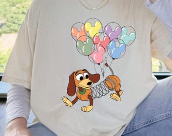 Può includere: T-shirt beige con un'illustrazione a cartoni animati di Slinky Dog di Toy Story. Slinky è raffigurato con palloncini colorati a forma di Topolino. Il design include elementi marroni, arancioni, gialli, rosa, verdi, viola e blu.