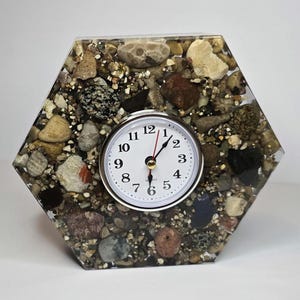 May include: A hexagonal desk clock featuring a clear resin body embedded with a variety of small, multi-colored natural stones and pebbles. The clock face is white with black Arabic numerals, black hour and minute hands, and a red second hand.