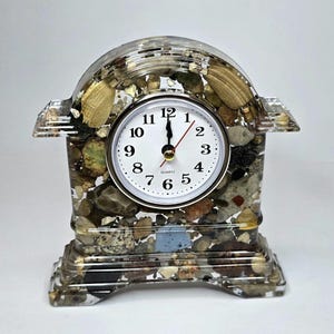 May include: A mantel clock with an arched top, encased in clear resin filled with various small, multi-colored natural stones. The clock face is white with black Arabic numerals, a silver-tone bezel, and black hour and minute hands.