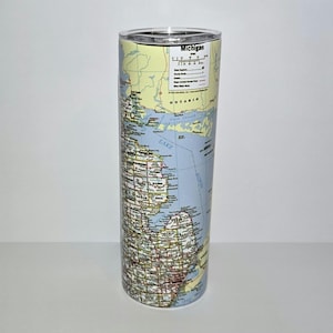 Puede incluir: Un vaso cilíndrico con un mapa de Michigan y sus alrededores. El mapa incluye el nombre del estado y una pequeña leyenda. El vaso tiene una tapa transparente y un interior blanco, con el diseño del mapa envolviendo el exterior.