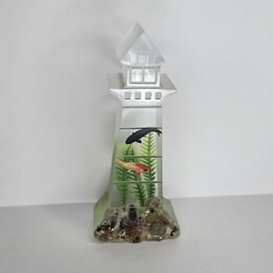May include: A clear, lighthouse-shaped decorative object featuring a black fish and a red and white fish swimming among green aquatic plants. The base is filled with small, textured gray and brown stones, and the top section resembles a lantern room.