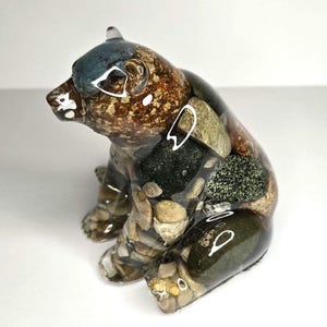 May include: A bear-shaped figurine crafted from clear resin embedded with a variety of smooth, multi-colored river stones and pebbles. The sculpture features a glossy, reflective surface that showcases natural earth tones including brown, gray, and green.