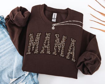 May include: A brown sweatshirt with the word "MAMA" in leopard print lettering. A silver necklace with a pendant is draped across the sweatshirt. The sweatshirt is folded on top of a pair of blue jeans.