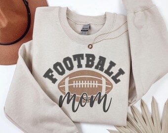 May include: A beige sweatshirt with the words "FOOTBALL MOM" in a combination of gray and brown. The word "FOOTBALL" is above a brown football graphic, and "mom" is below. A brown hat and gold necklace are visible.