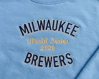 Può includere: Una felpa girocollo azzurra con testo ricamato. La parola MILWAUKEE è in blu scuro, seguita dal testo dorato World Series 2026 e dalla parola BREWERS in blu scuro in basso. Il capo presenta un colletto a coste.