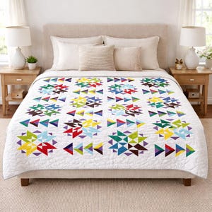 May include: A white quilt with a geometric pattern of colorful triangles in red, yellow, green, blue, and purple. The quilt is spread over a bed with neutral-toned bedding and wooden nightstands.