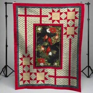 Könnte beinhalten: Eine gesteppte Decke mit einem zentralen Feld mit roten Kardinälen auf Zweigen mit Weihnachtssternen. Die Decke hat einen roten Rand, beige Sternakzente und einen cremefarbenen Hintergrund mit kleinen, sich wiederholenden Mustern.