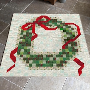 Op de afbeelding: Een gewatteerd kerstkransontwerp op een crèmekleurige achtergrond. De krans is gemaakt van groene en rode vierkanten, met een grote rode strik. De quilt is ongeveer 76 cm vierkant.