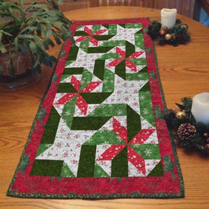 Peut inclure: Une nappe de table rectangulaire de Noël avec un motif patchwork. La nappe présente une bordure rouge avec un motif de poinsettia et un motif central de formes géométriques vertes, blanches et rouges. Le motif comprend des flocons de neige et des baies de houx.