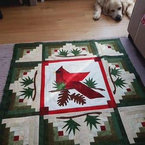 May include: A handmade quilt featuring a central design of a red cardinal bird perched on a branch with green foliage. The quilt has a square pattern with green and white blocks, and a red border. A golden retriever dog is in the background.