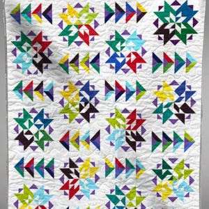 May include: A white quilt with a geometric pattern of colorful triangles and starburst shapes. The quilt features a repeating design in shades of red, yellow, green, blue, and purple. The edges are finished with a black and white striped border.