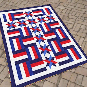 May include: A rectangular quilt with a patriotic design. The quilt features a white background with red and blue rectangular blocks and star-shaped accents. The border is a dark blue, and the quilt is placed on a stone surface.