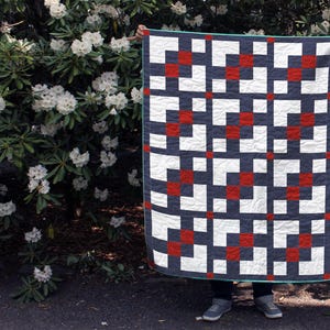 May include: A handmade quilt with a geometric pattern of navy blue, white, and red squares. The quilt is held up against a backdrop of green foliage and white flowers. The quilt has a teal border.