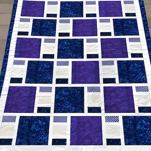 May include: A handmade quilt with a geometric pattern. The quilt features large squares of deep blue and purple, interspersed with white and light blue rectangles. The quilt is bordered with a dark blue fabric.