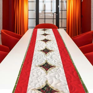 May include: A red and white quilted table runner with a central pattern of diamond shapes filled with red and green floral designs. The runner has a red border and is placed on a white table.