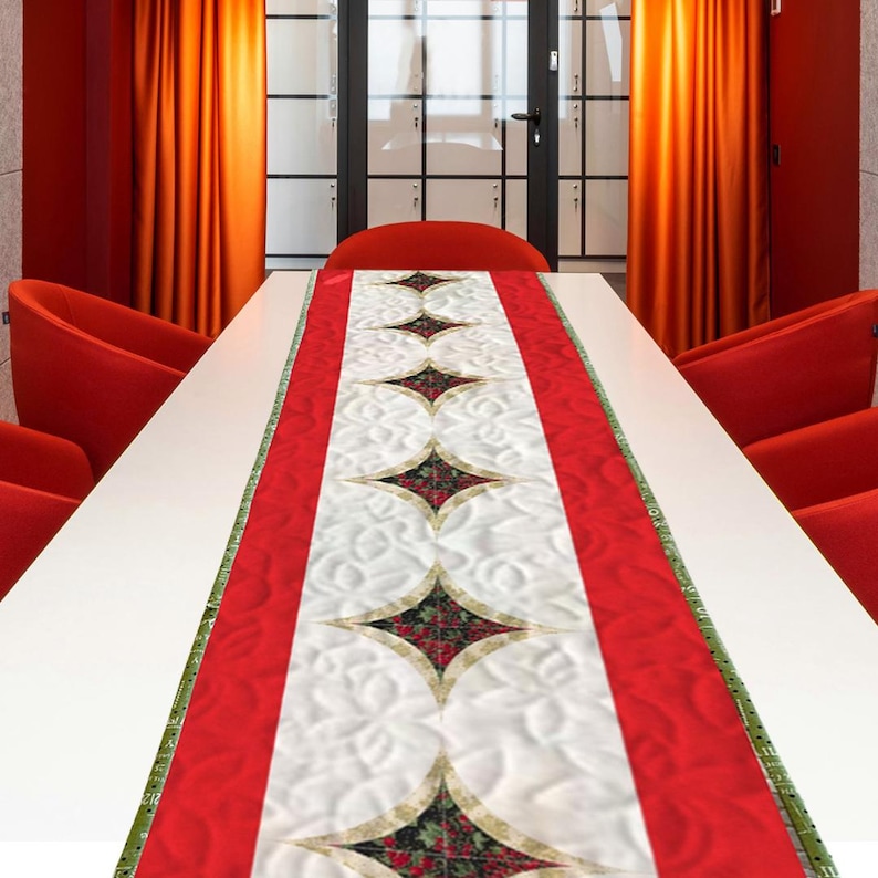 K&ouml;nnte beinhalten: Ein rot-wei&szlig; gesteppter Tischl&auml;ufer mit einem zentralen Muster aus rautenf&ouml;rmigen Formen, die mit roten und gr&uuml;nen floralen Designs gef&uuml;llt sind. Der L&auml;ufer hat einen roten Rand und liegt auf einem wei&szlig;en Tisch.