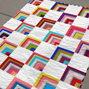May include: A colorful quilt with a geometric design. The quilt features white squares with vibrant, layered, L-shaped patterns in shades of red, pink, yellow, blue, and green. The quilt is made of fabric and has a textured surface.
