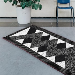May include: A rectangular quilt with a geometric pattern in black, white, and gray. The design features triangles and diamond shapes, with a dotted border and a burgundy edge. The quilt is placed on a gray tiled surface.