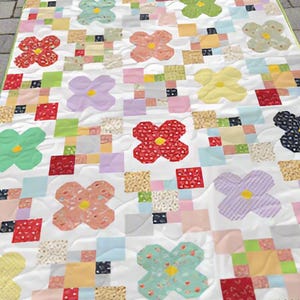 May include: A handmade quilt with a repeating pattern of colourful flower shapes and patchwork squares. The flowers are in shades of red, pink, yellow, green, blue, and purple. The quilt has a white background and a green border.