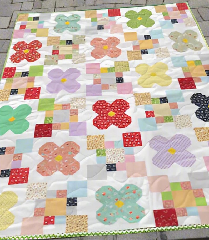May include: A handmade quilt featuring a repeating pattern of colorful flower shapes and patchwork squares. The flowers are in shades of red, pink, yellow, green, blue, and purple. The quilt has a white background and a green border.