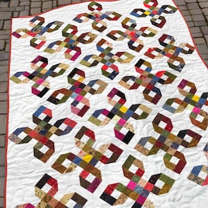 May include: A handmade quilt with a white background and a repeating pattern of colorful, interlocking shapes. The shapes are made of various fabric scraps in shades of blue, green, red, and brown. The quilt has a red border.