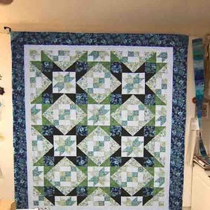 May include: A large, handmade quilt with a star pattern. The quilt features a white background with squares of green, blue, and black fabric. The border is a dark blue floral print. The quilt is hanging on a wall.