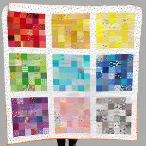 May include: A colorful patchwork quilt with a white dotted border and orange trim. The quilt is divided into nine squares, each a different color: red, yellow, green, blue, purple, pink, and gray. The squares are made of smaller fabric pieces.