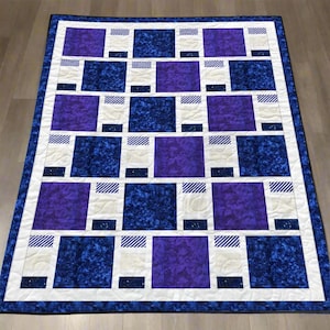 May include: A handmade quilt with a geometric pattern. The quilt features alternating squares of deep blue, purple, and white, with striped and speckled accents. The quilt is bordered with a white and blue frame.