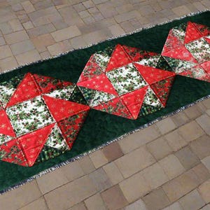 Pode incluir: Um caminho de mesa de Natal acolchoado com três grandes desenhos em forma de estrela. As estrelas são feitas de tecido vermelho com padrões de flocos de neve brancos e folhas de azevinho verdes. O caminho de mesa tem um fundo verde escuro e uma borda com franjas.
